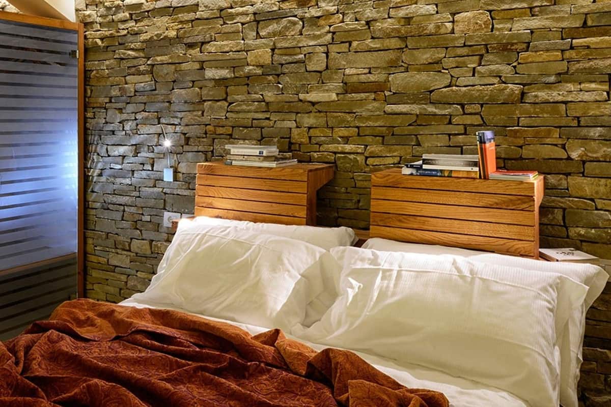 A bedroom with a stone wall, a glass shower enclosure, wooden headboards, and a bedside table with books.
