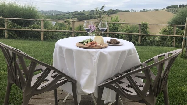 Una mesa redonda con mantel blanco, rodeada de sillas metálicas, ubicada al aire libre con vistas a colinas ondulantes y una terraza de madera.