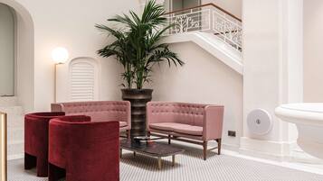 A modern interior with a pink sofa set, a red ottoman, a potted plant, and a staircase with white railings.