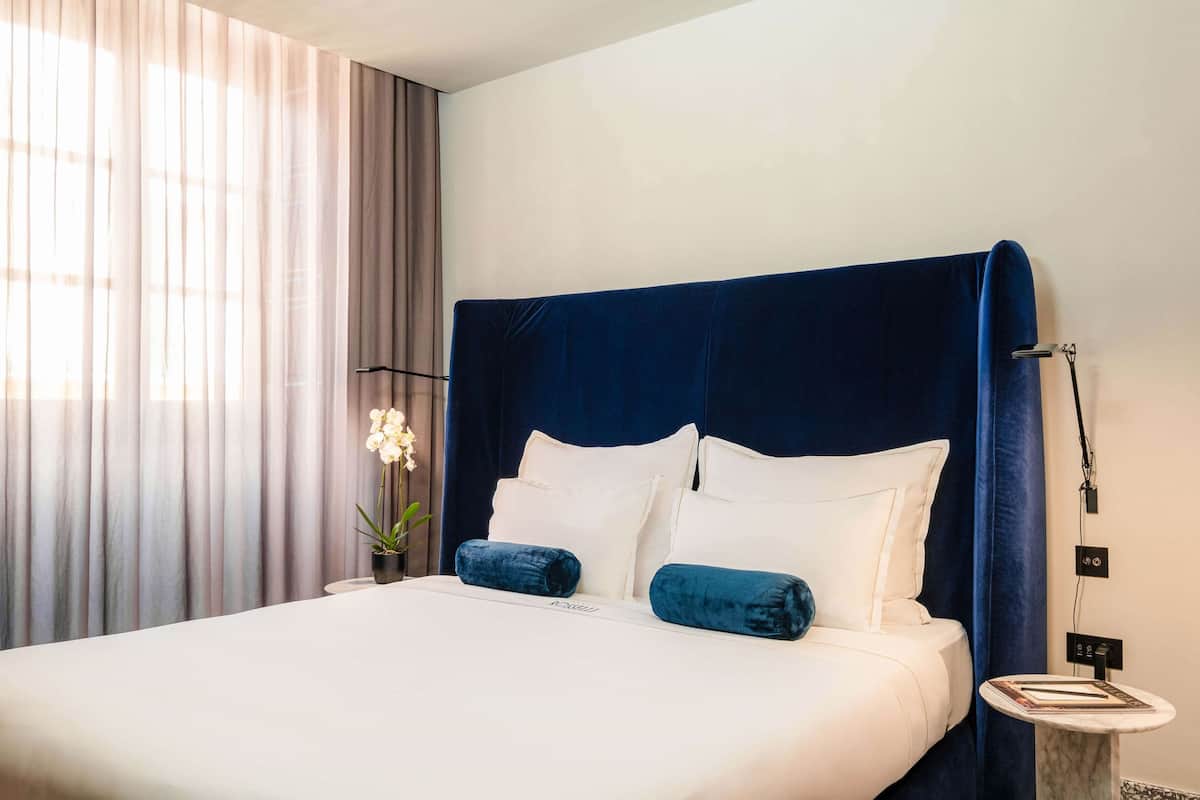 A neatly made bed with white and blue bedding, a bedside table with a lamp, and a window with sheer curtains.