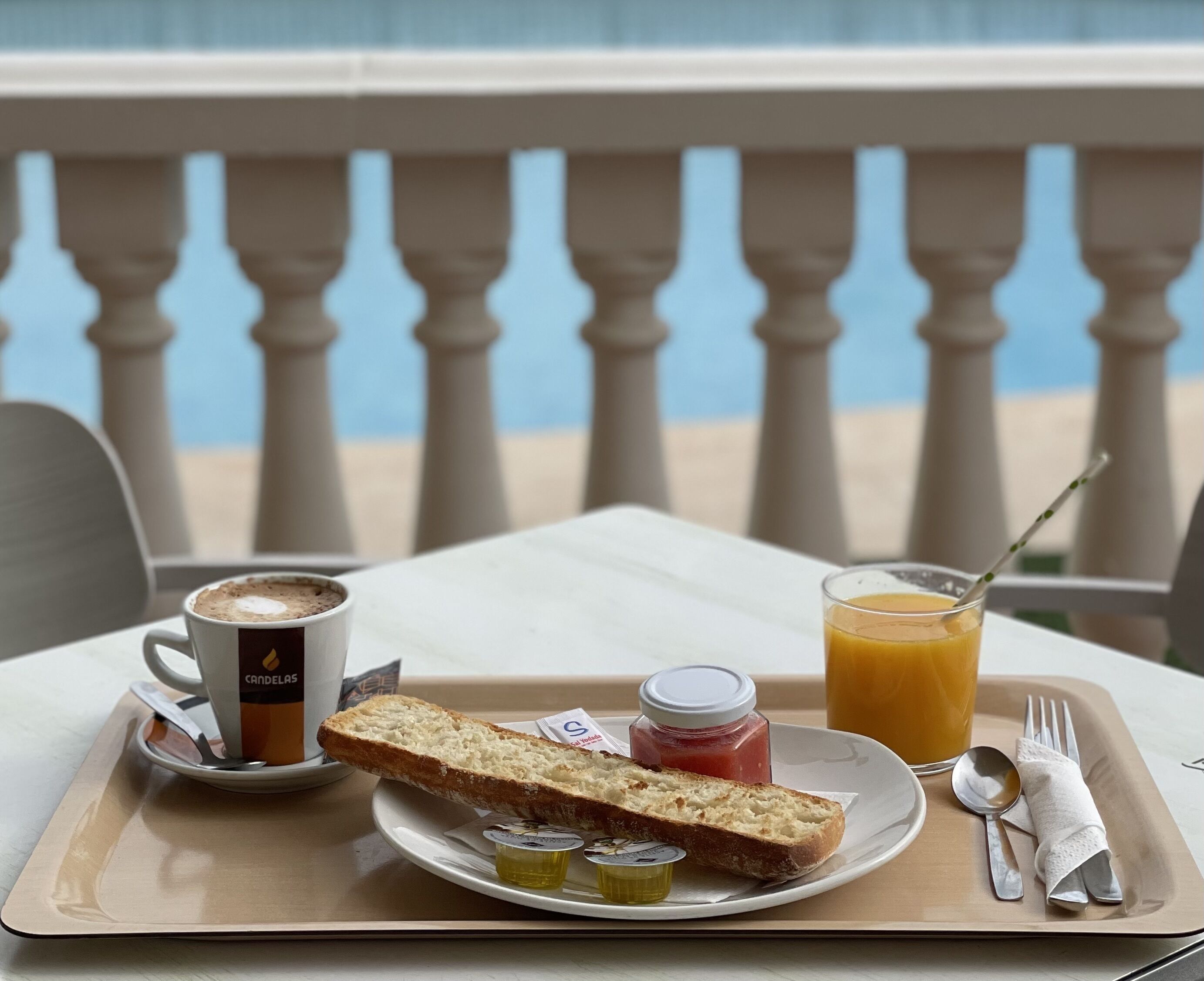 A breakfast tray with bread, jam, butter, a cup of coffee, and a glass of orange juice on a balcony with a railing.
