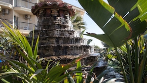 Una fuente de piedra con agua en cascada, rodeada de exuberante vegetación y un edificio con balcones al fondo.