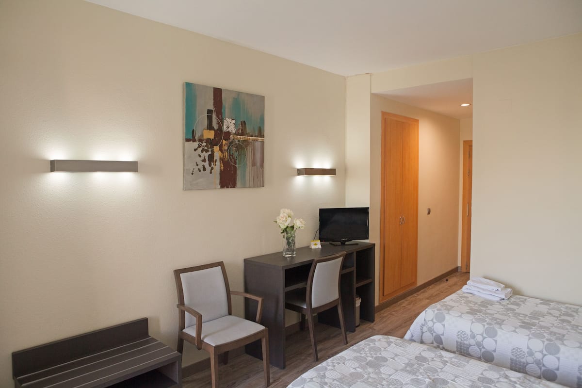 Quarto de hotel com cama, escrivaninha com cadeira, televisão e um quadro na parede.