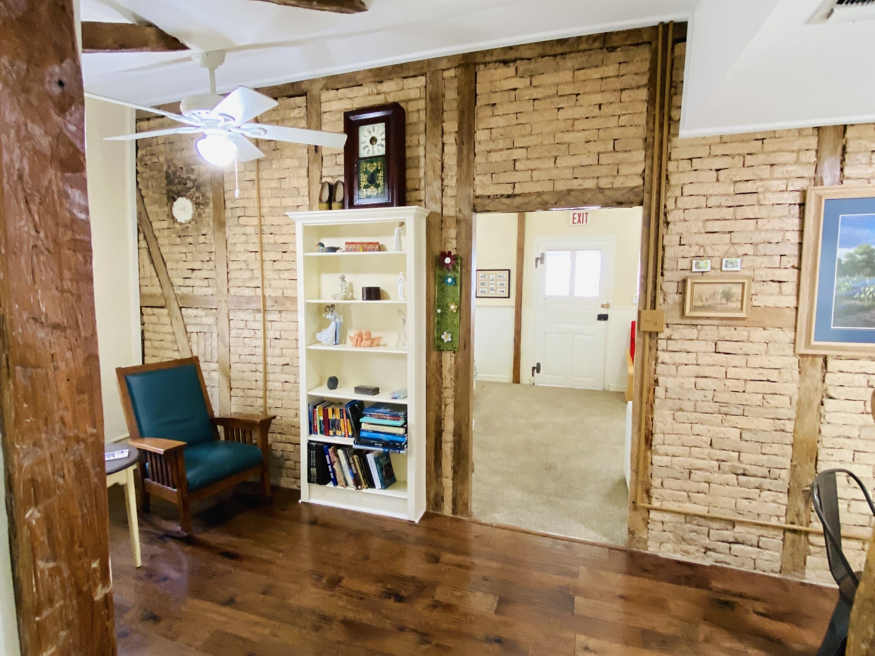 Une pièce avec un mur en briques, une bibliothèque blanche, une chaise en bois et un ventilateur de plafond.