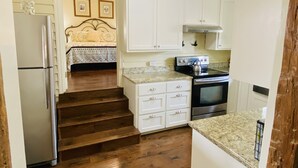 A compact kitchen with white cabinets, a refrigerator, a stove, and a microwave. There are wooden stairs leading to a bedroom with a bed and a headboard.