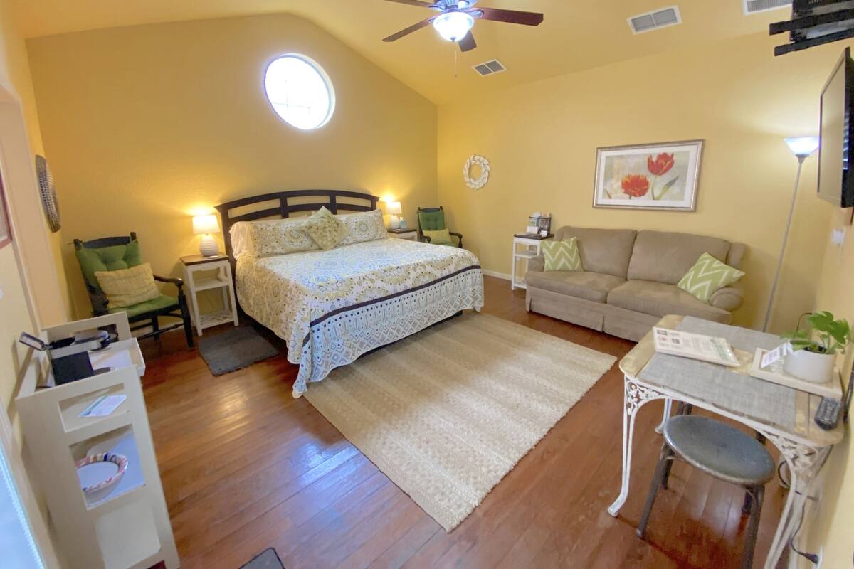 Ein Schlafzimmer mit einem Bett, einem Schreibtisch, einem Sessel, einer Couch, einem Deckenventilator, einer Wanduhr und einem Fenster mit Tageslicht.