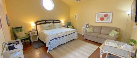 A bedroom with a bed, a desk, a chair, a sofa, a ceiling fan, a wall clock, and a window with natural light.