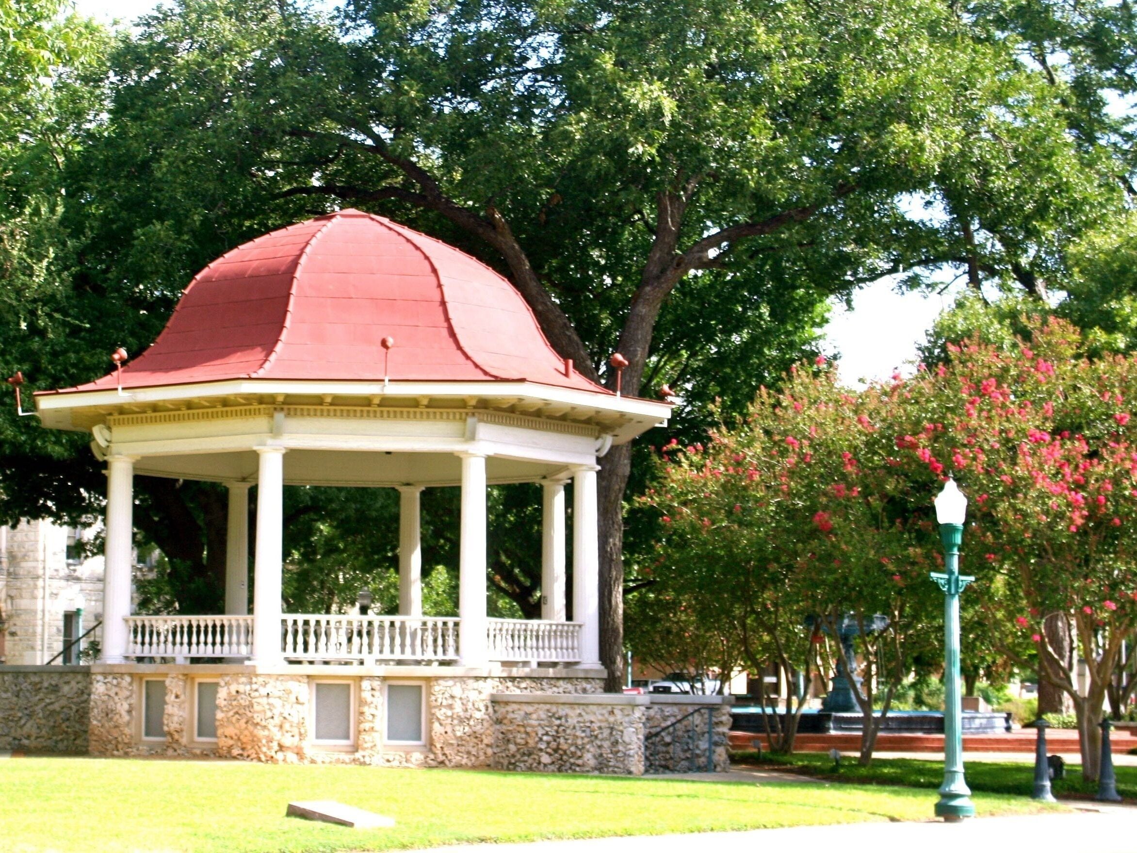 Un kiosque avec un toit rouge et des colonnes blanches, entouré d’arbres et d’un mur de pierre.