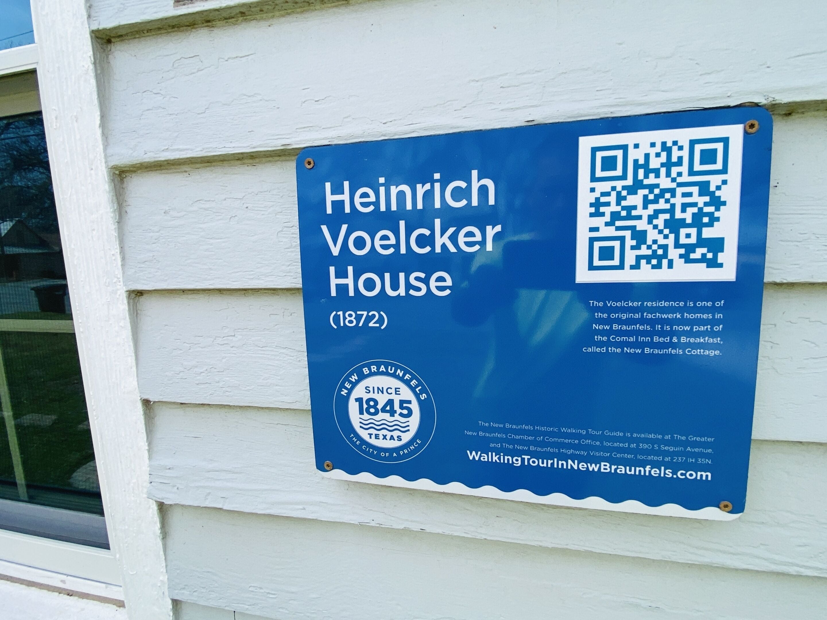 A blue sign on a white building wall. The sign reads "Heinrich Voelcker House (1872)" and includes a QR code.