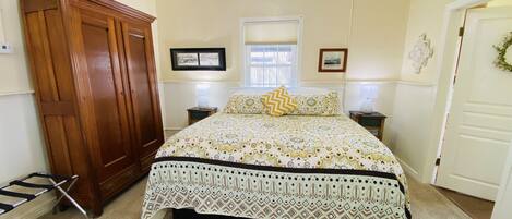 Un dormitorio con un armario de madera grande, una cama con ropa de cama estampada, dos mesitas de noche con lámparas y una ventana con persianas.