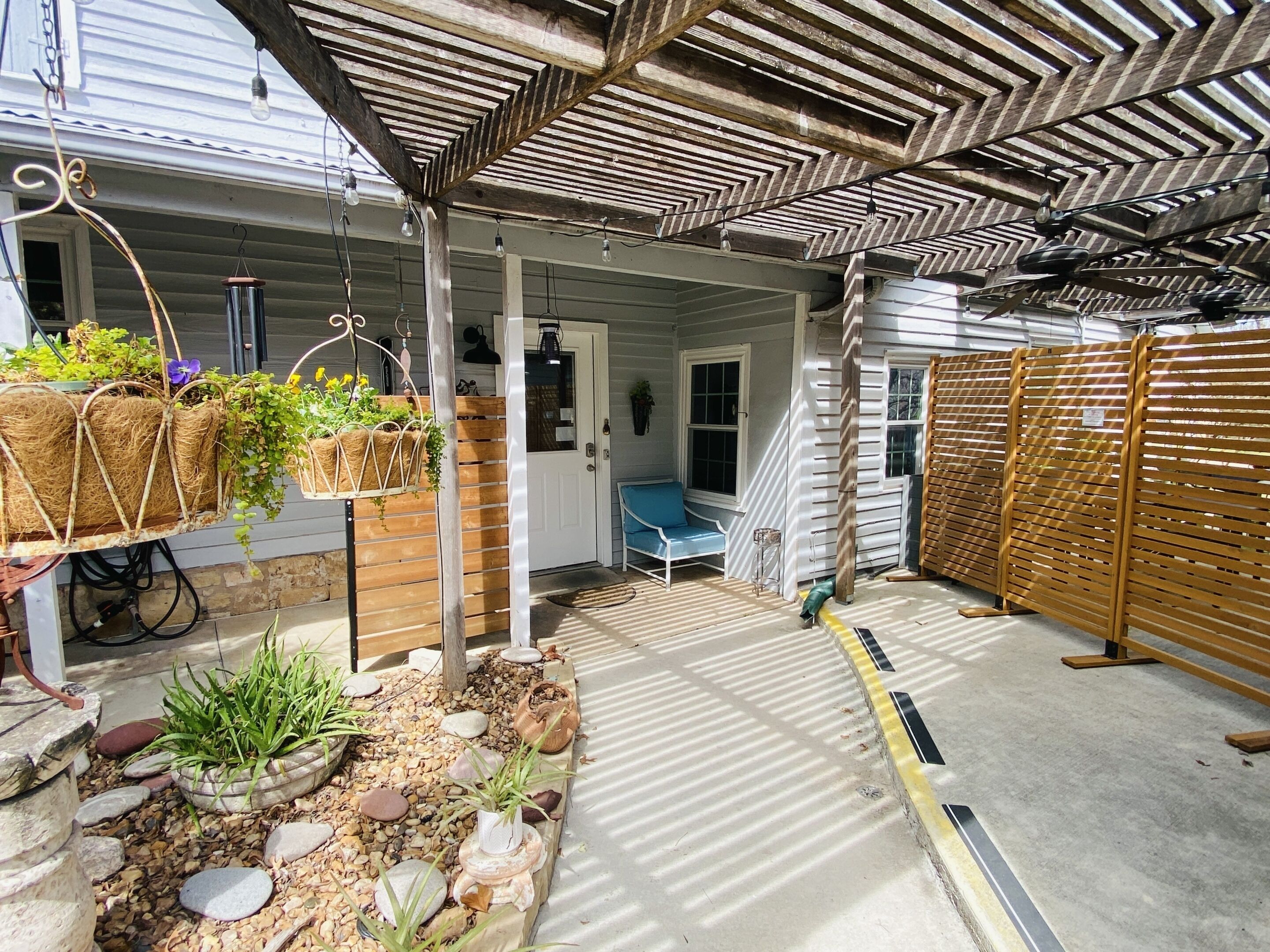 A covered outdoor area with a wooden pergola, hanging planters with greenery, a bench, and a wooden fence.