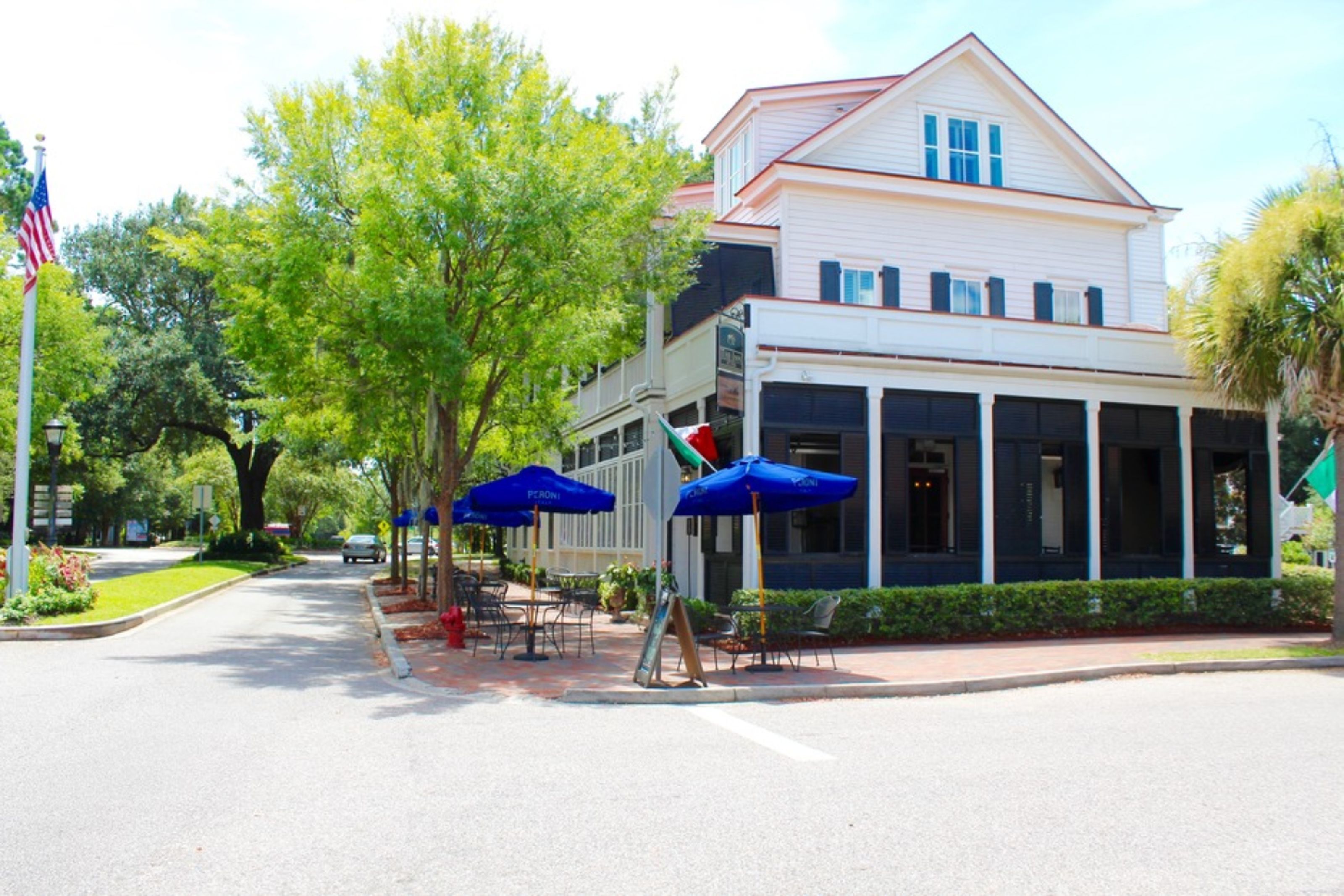 A two-story building with a white facade and a red roof. There are outdoor seating areas with blue umbrellas and a flagpole with an American flag.