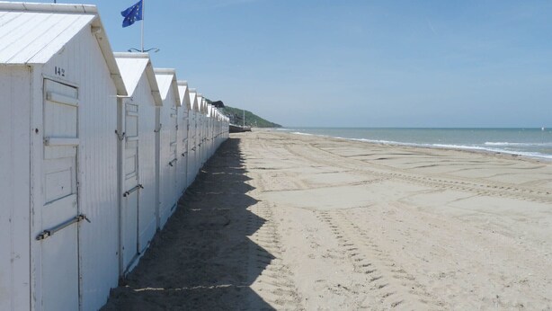 Una hilera de cabañas blancas frente a la playa, cielo despejado y arena.