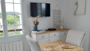 Un comedor con mesa de madera y sillas de mimbre, televisor montado en la pared y ventana con vistas a zonas verdes.