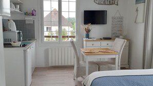 Habitación compacta y bien iluminada con cama, kitchenette pequeño, zona de comedor con mesa y sillas, y televisor montado en la pared.