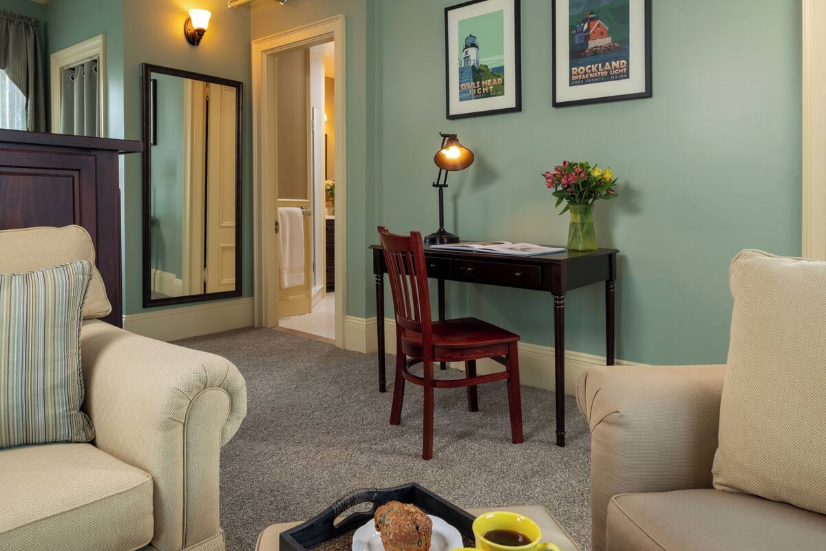 A cozy living room with a sofa, a chair, a desk with a lamp, a tray with a muffin and a cup, and framed pictures on the wall.