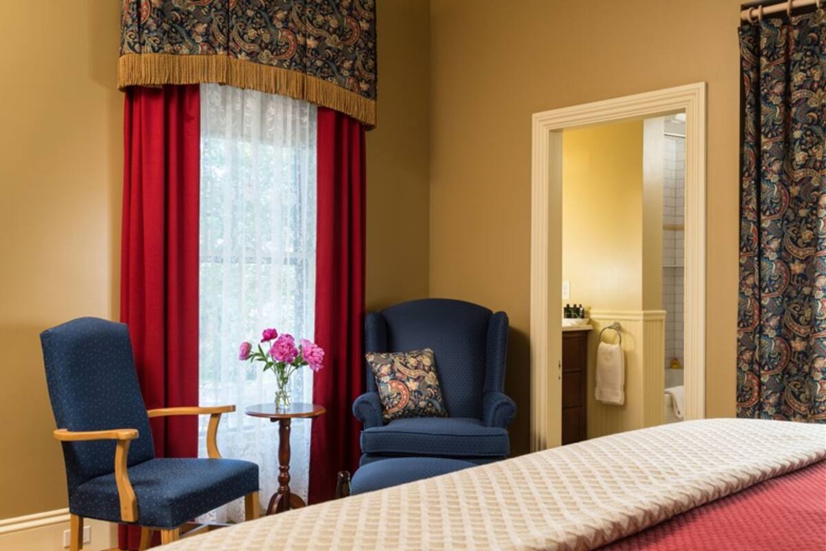 A hotel room with a bed, two armchairs, a small table with a vase of flowers, and a window with red curtains.
