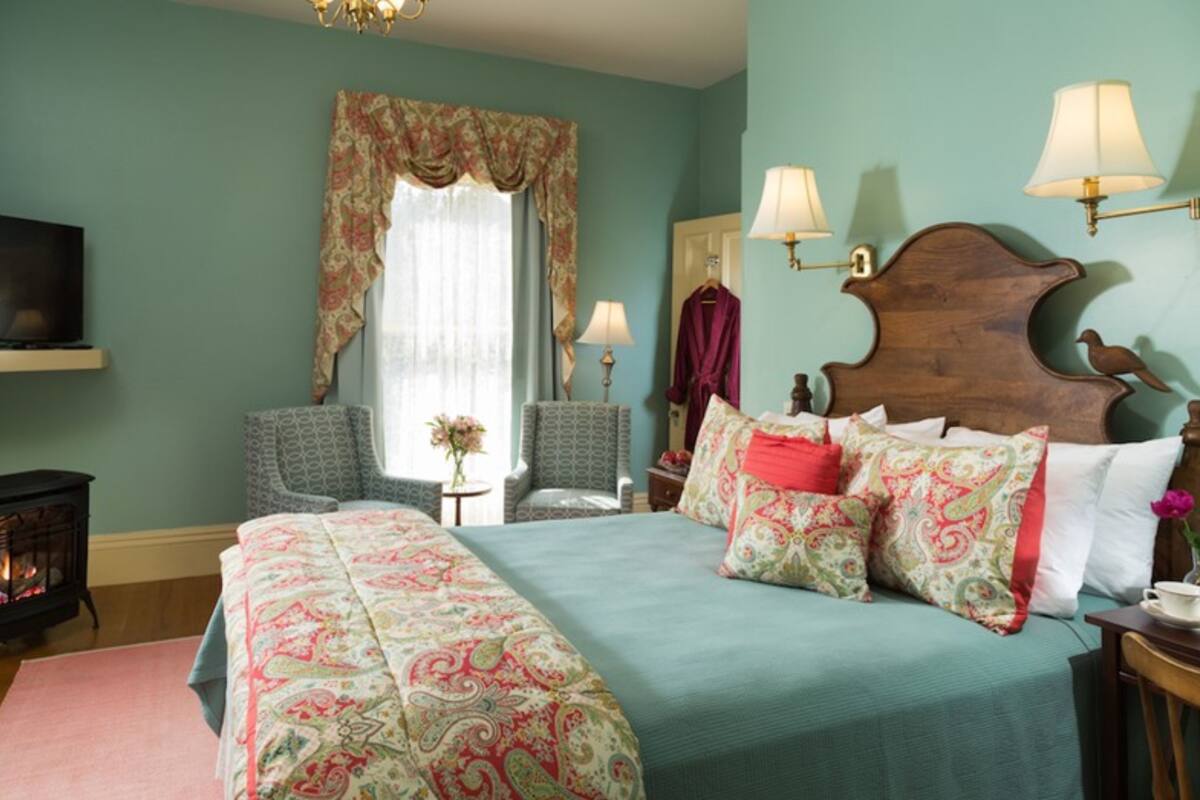 A bedroom with a large bed, two chairs, a television, a fireplace, and wall-mounted lamps.