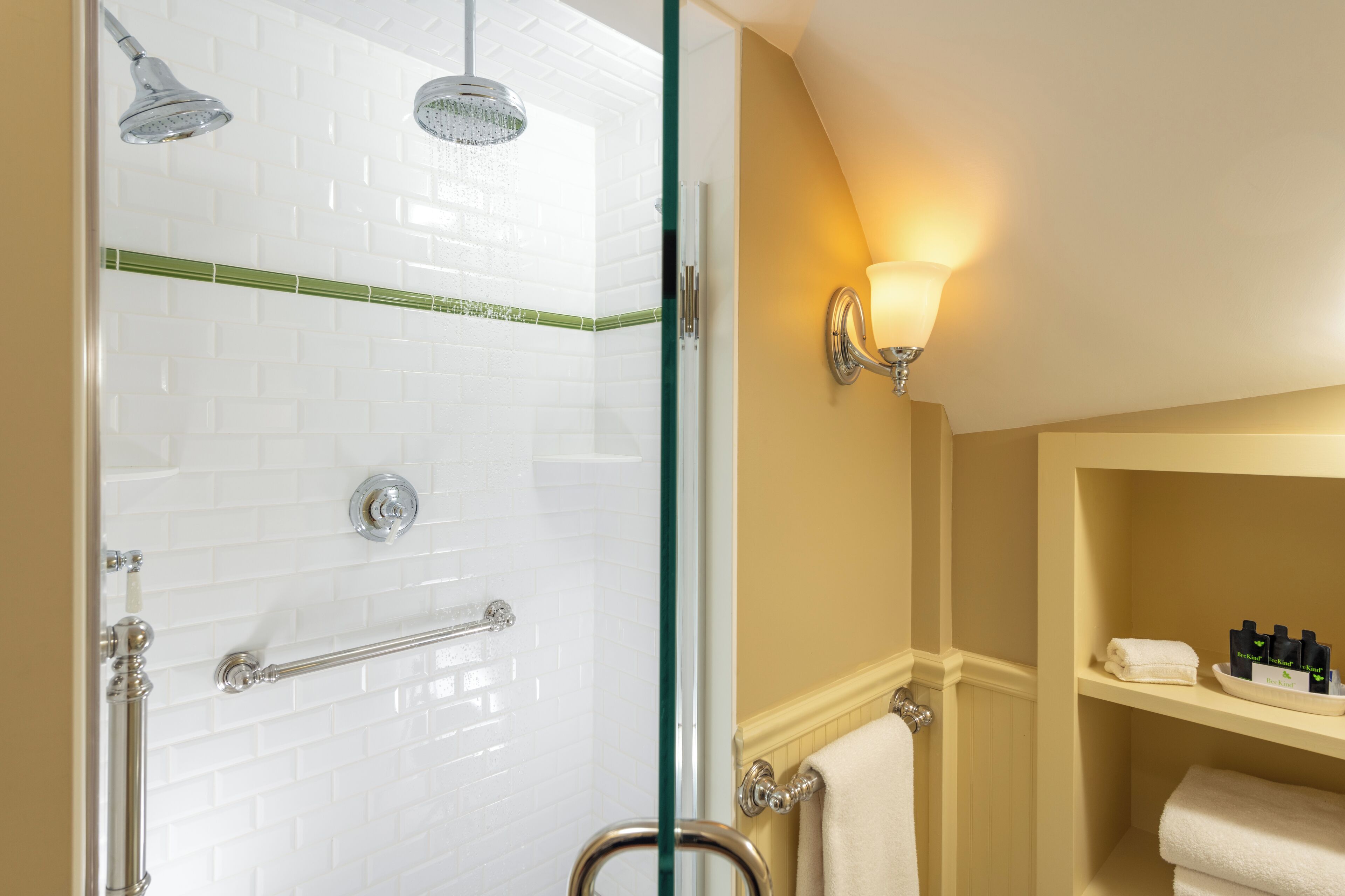A bathroom with white tiled walls, a glass shower enclosure, a towel rack, and a shelf with toiletries.