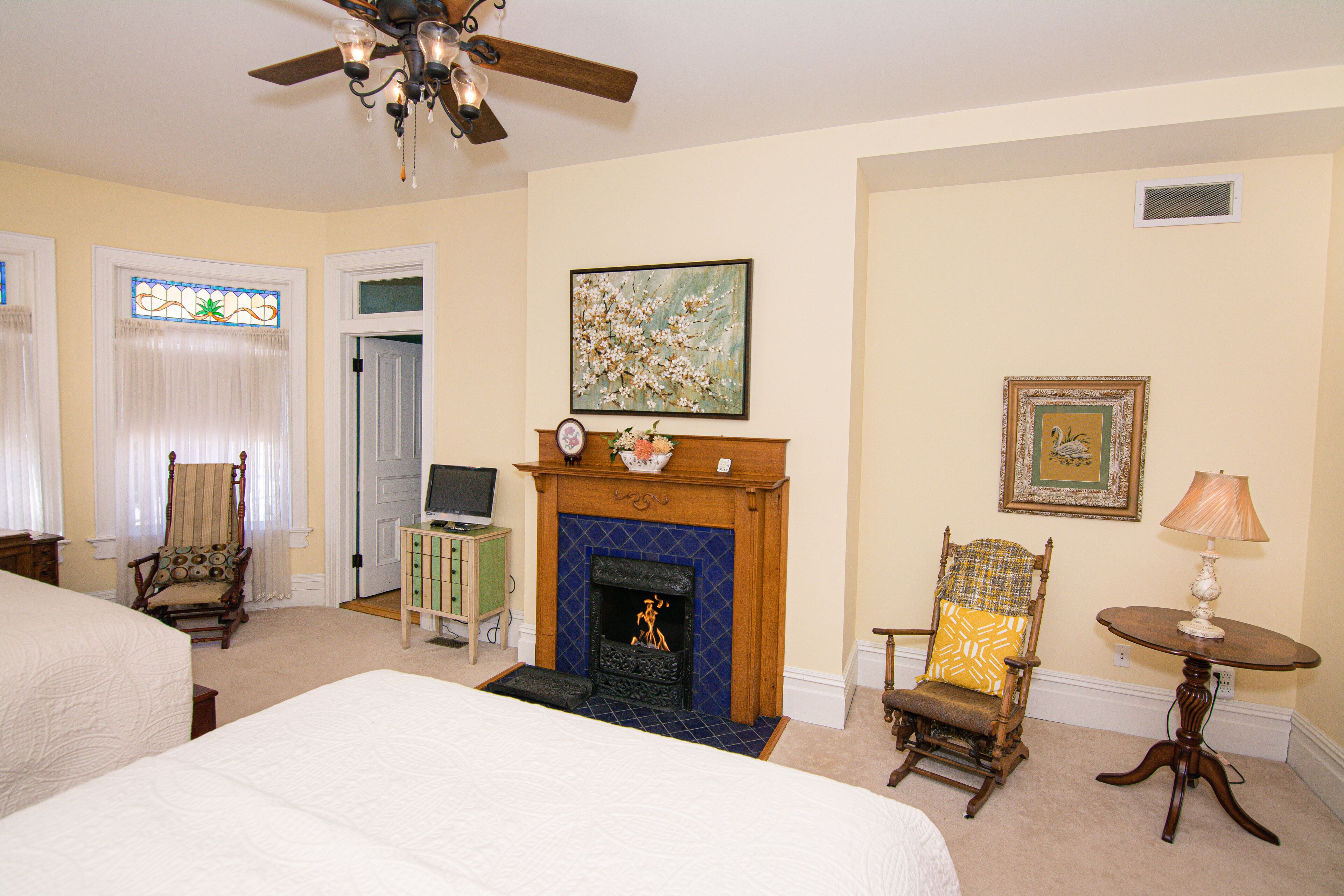 A bedroom with a fireplace, a bed, a chair, a table, and a ceiling fan.