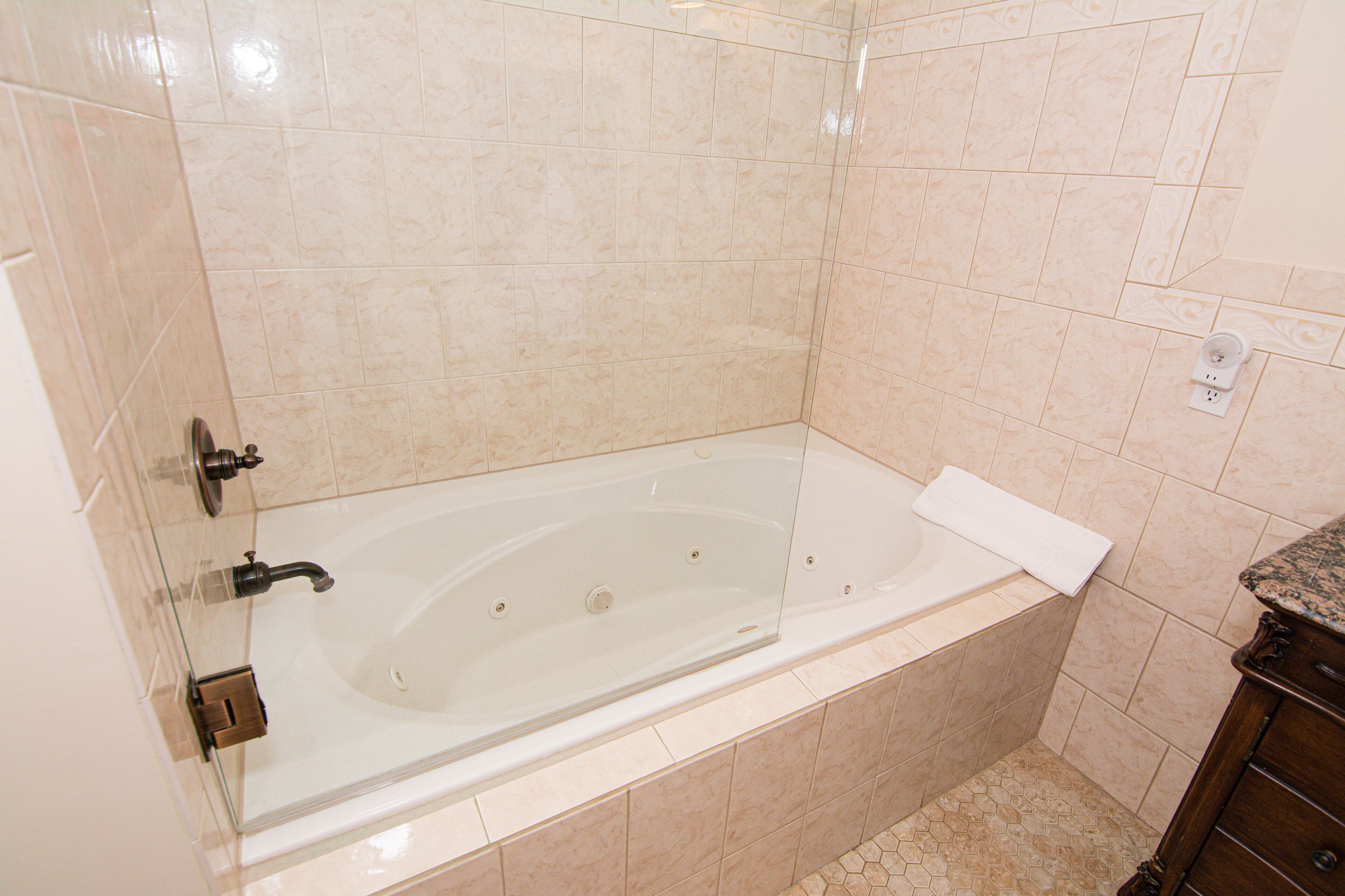 Un baño moderno con bañera de hidromasaje con mampara de vidrio, azulejos color beige y un mueble de tocador de madera.