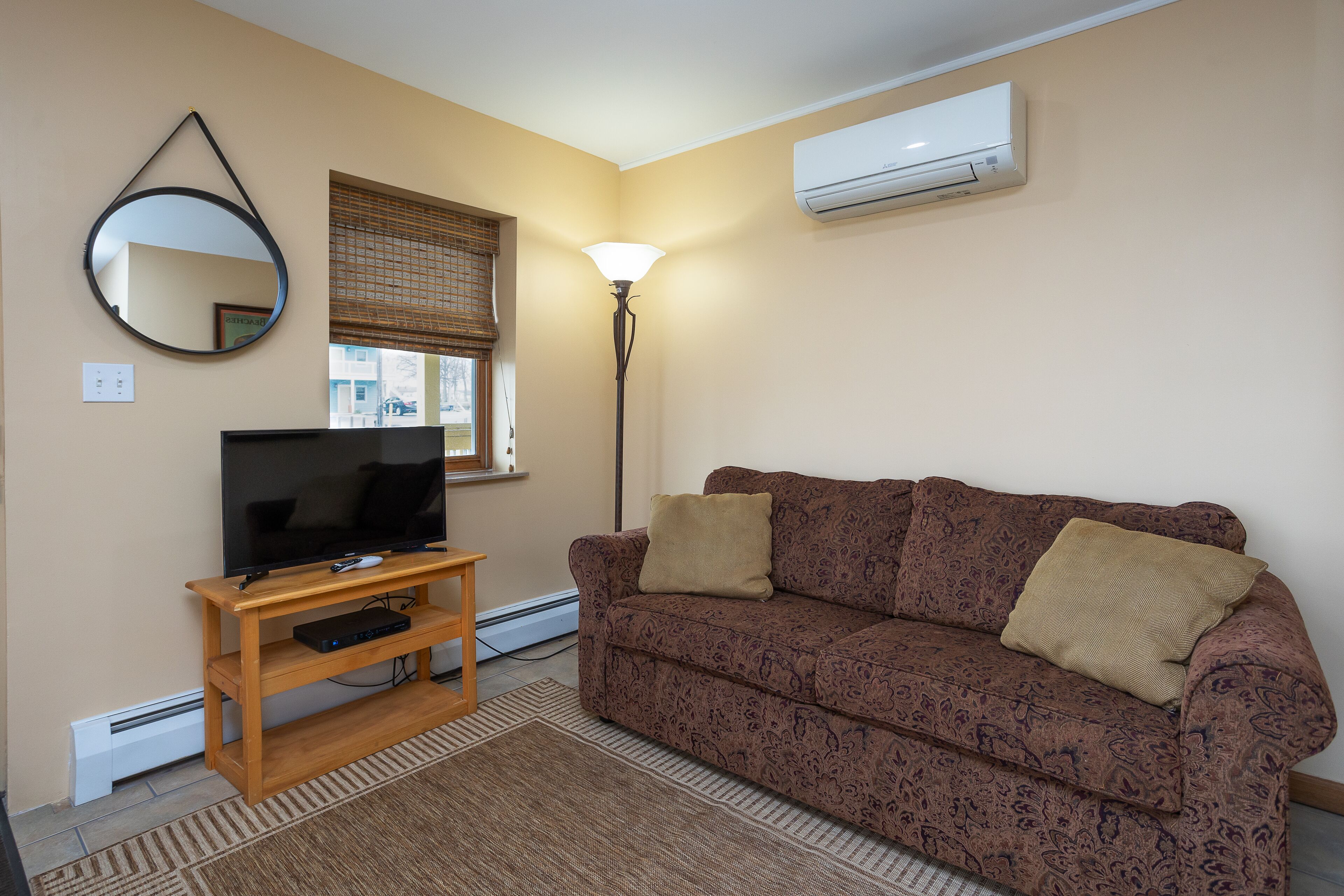 Ein Wohnzimmer mit einer Couch, einem Fernseher auf einem Holzboden, einer Lampe und einer Klimaanlage an der Wand.