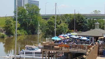 A riverside dock with boats, a wooden pier with tables and umbrellas, and a bridge in the background.