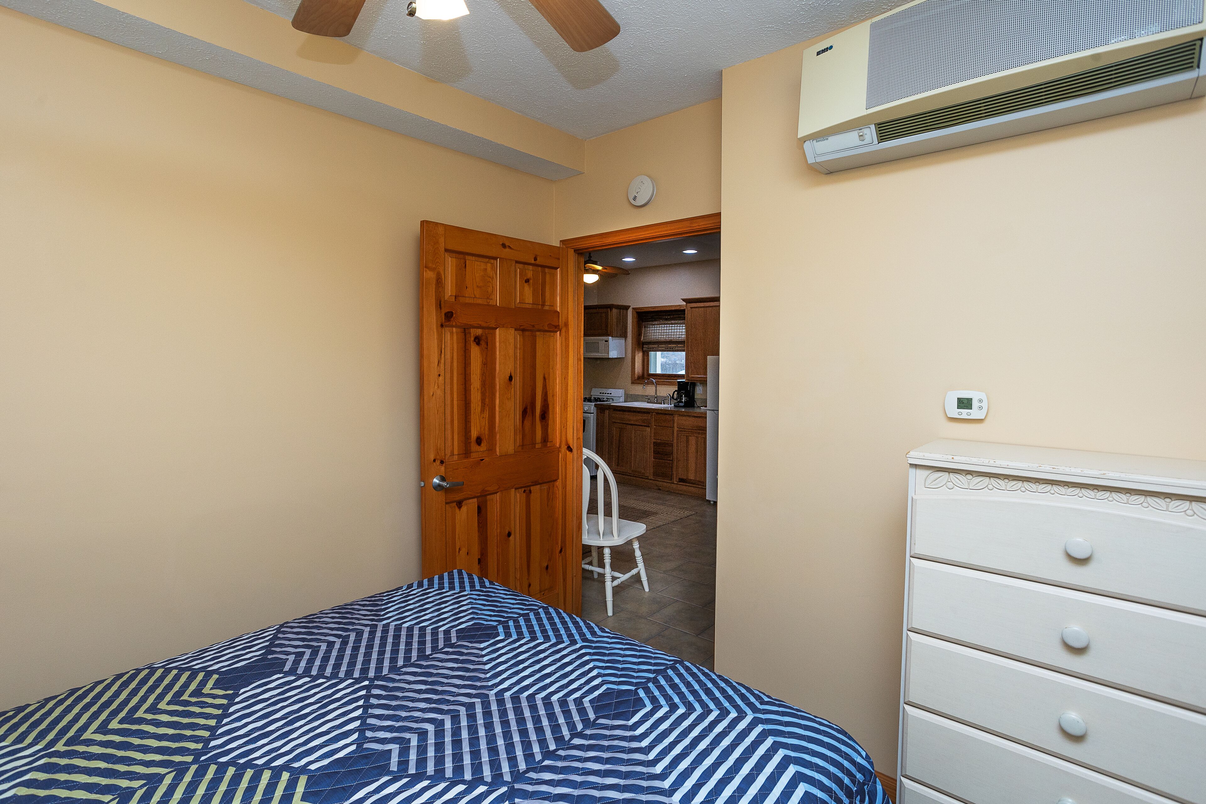 A bedroom with a bed, a wooden door, a white dresser, and a kitchenette in the background.