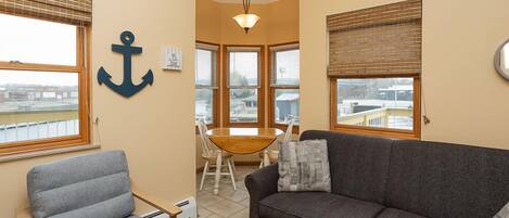 A cozy living room with a gray sofa, a gray armchair, and a wooden dining table with chairs. There is a large window with blinds, a ceiling light, and a wall-mounted anchor decoration.