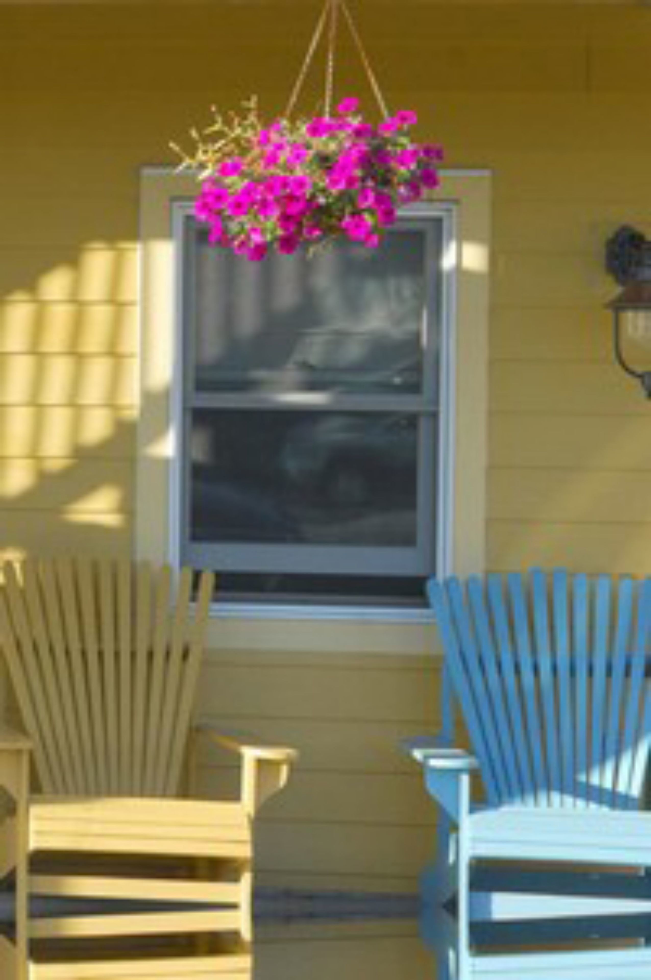 Une fenêtre ornée d’un pot de fleurs suspendu, deux fauteuils Adirondack et un luminaire fixé au mur.