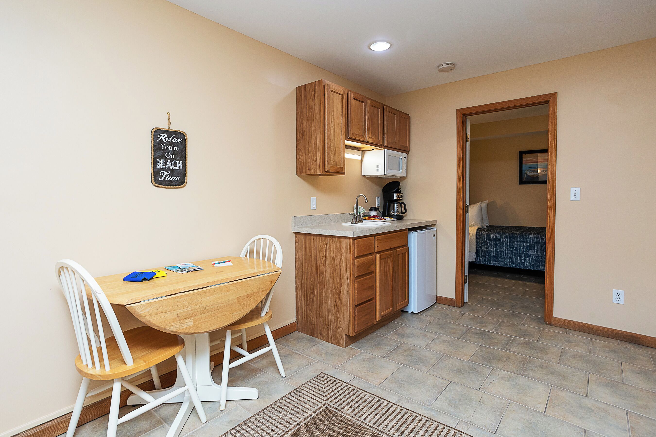 A compact kitchen with wooden cabinets, a microwave, and a coffee maker. There is a dining table with chairs and a rug on the floor.