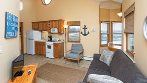 A compact living space with a kitchenette, a gray sofa, a wooden table, and a view of the water through the windows. - Bridge Inn (Michigan City)