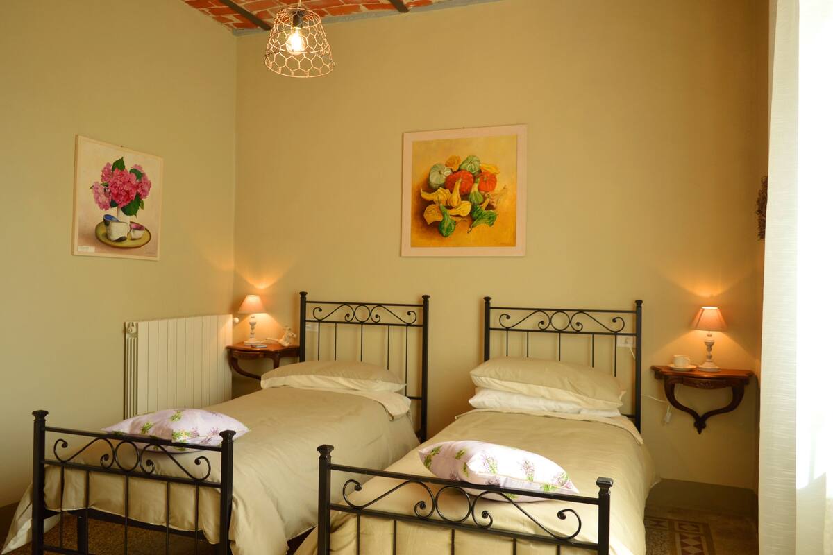 Una habitación con dos camas individuales, cada una con mesita de noche y lámpara. Hay dos cuadros en la pared, uno de flores y otro de verduras. La habitación tiene un diseño sencillo y funcional con un radiador en la pared.