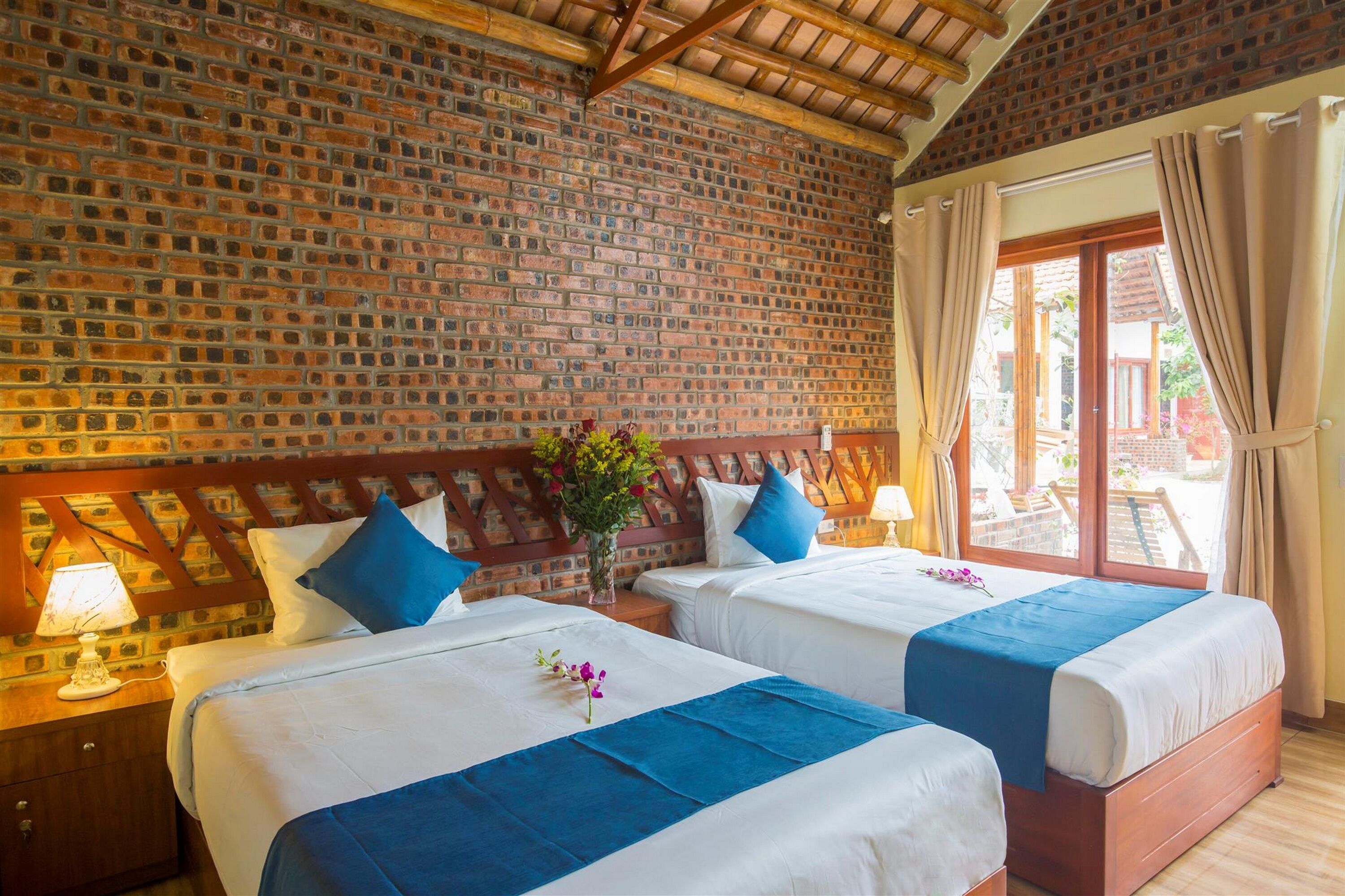 A hotel room with two beds, a brick wall, wooden ceiling beams, and a window with curtains.