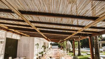 Un patio al aire libre con mesas y sillas, una sección cubierta con techo de bambú y una piscina al fondo.
