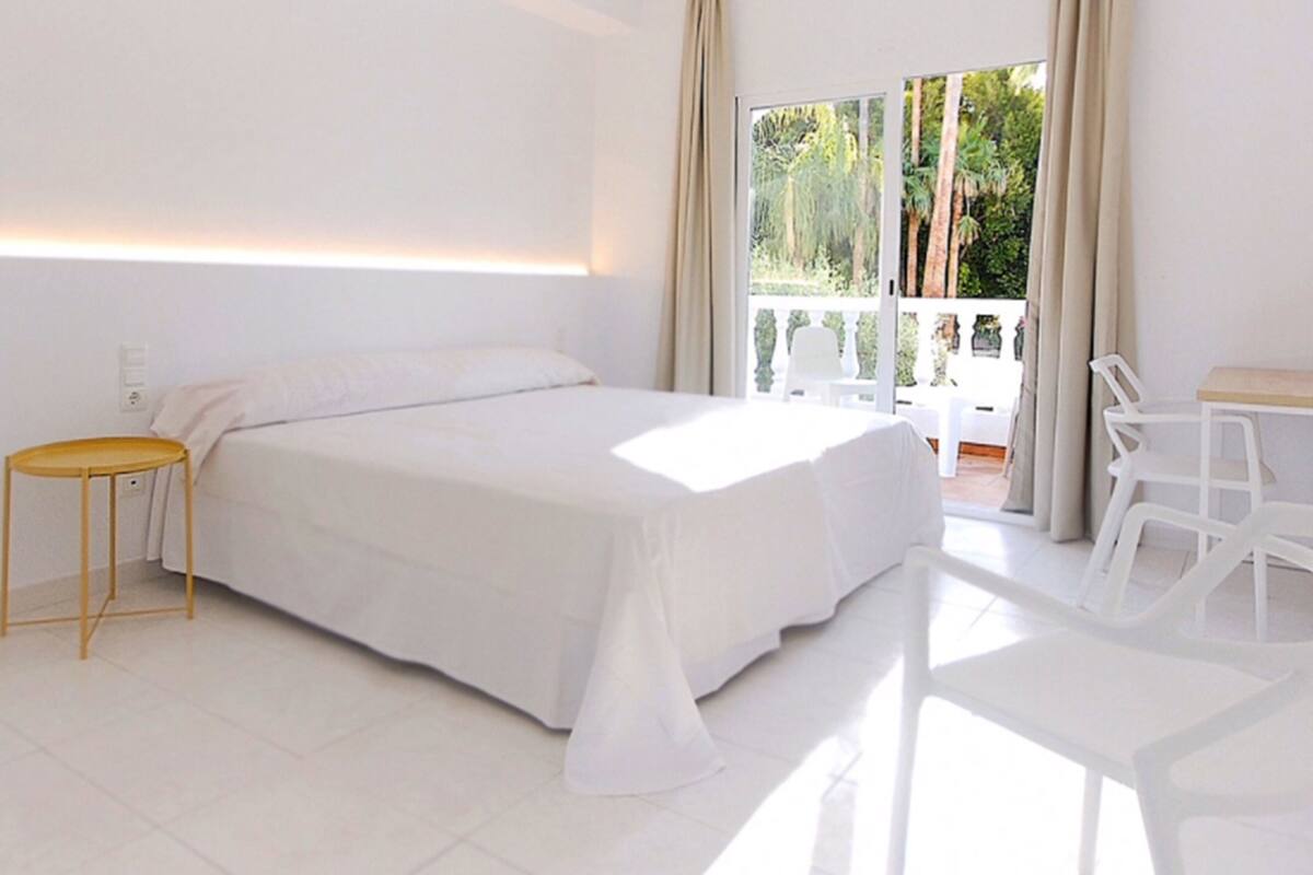 A modern bedroom with a large bed, a small side table, and a balcony with white chairs.