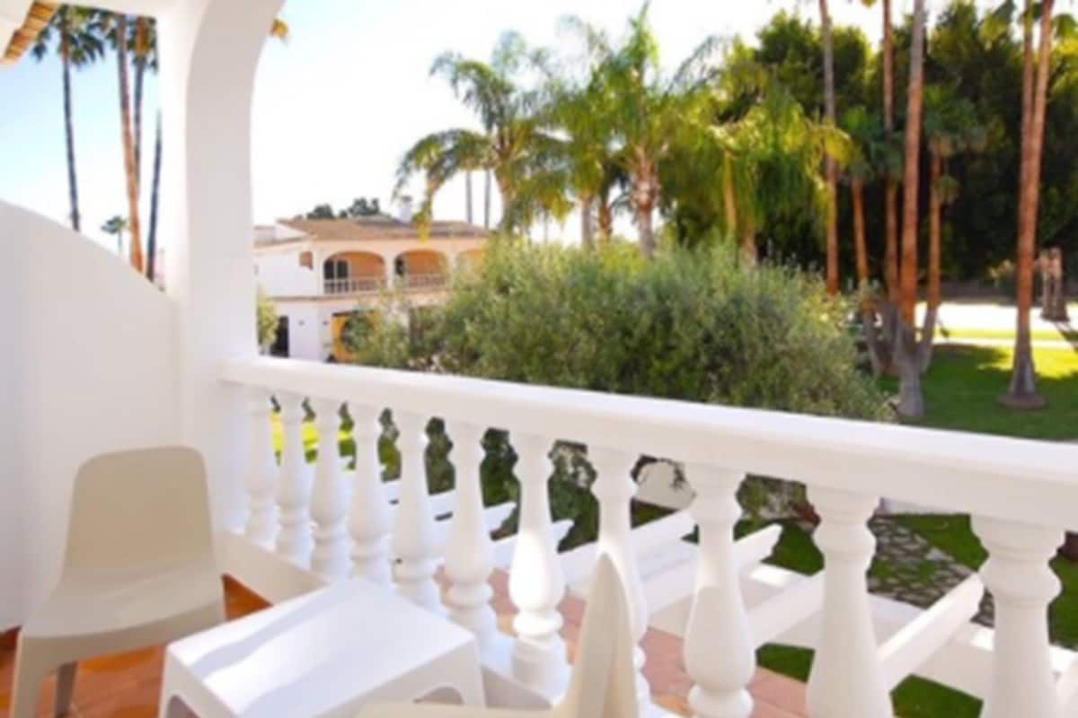 A balcony with white chairs and a table, overlooking a garden with palm trees and a building in the background.