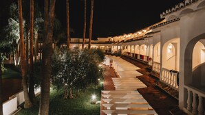 Vista nocturna de un pasillo de hotel con palmeras y arcos iluminados.