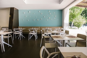 A modern dining area with white chairs and tables, a teal wall with bird decorations, and a view of greenery outside. - Tu&Me Resort - Adults Only (Gandia)