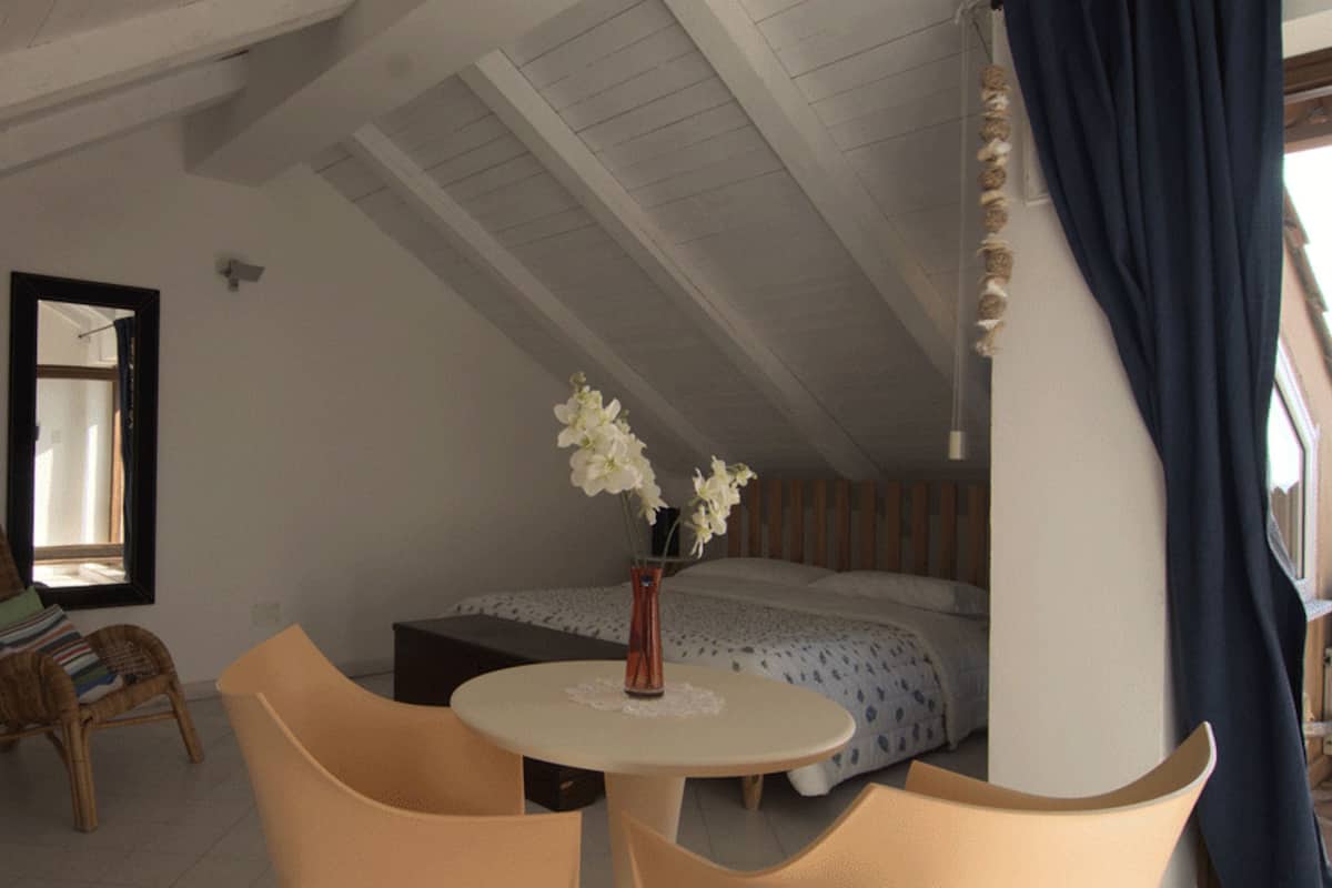 Une chambre à coucher avec un lit, une petite table sur laquelle est posé un vase de fleurs, une chaise et une fenêtre.