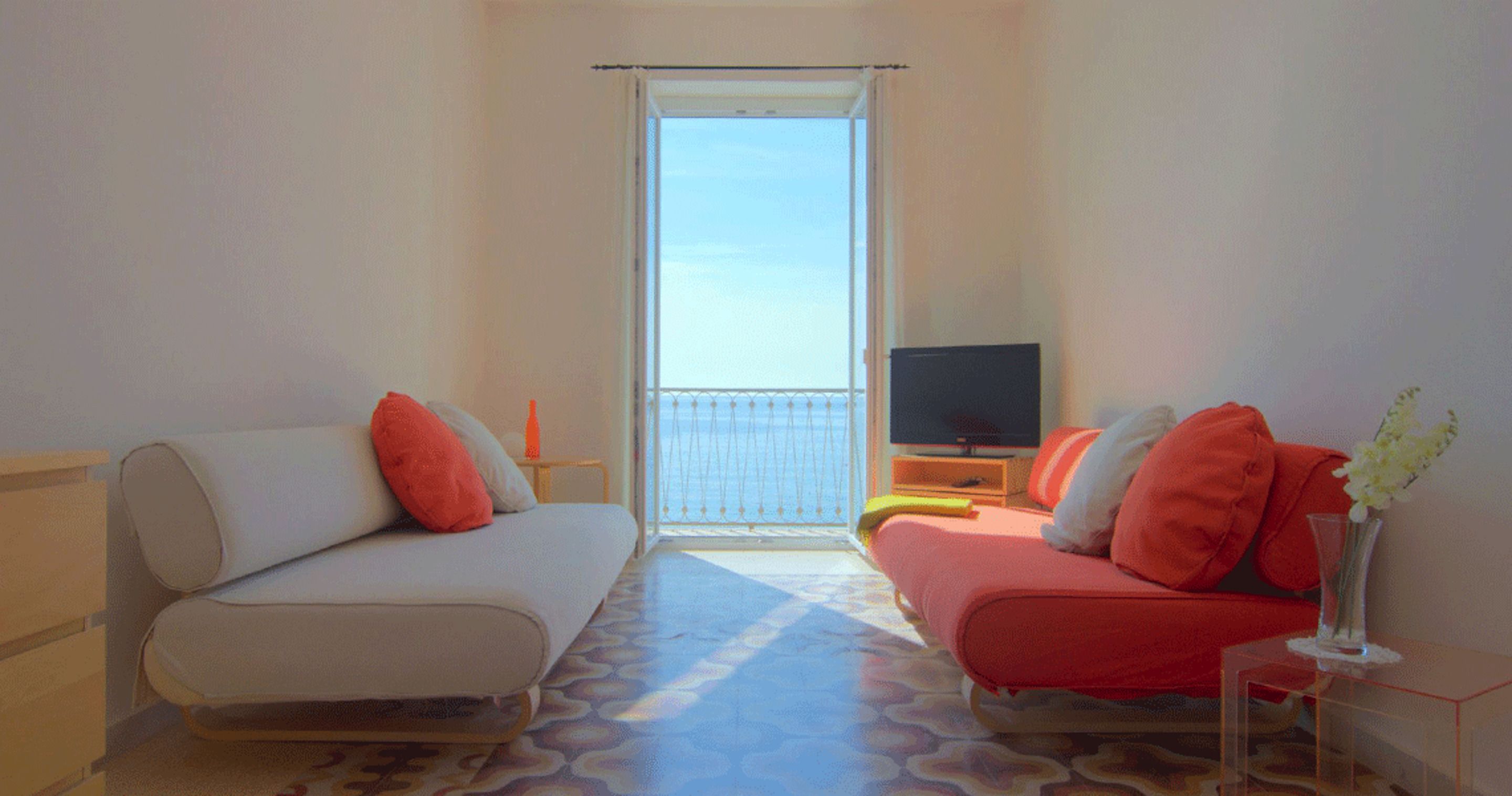 Quarto com sofá e poltrona de frente para uma varanda, com televisão sobre um móvel.