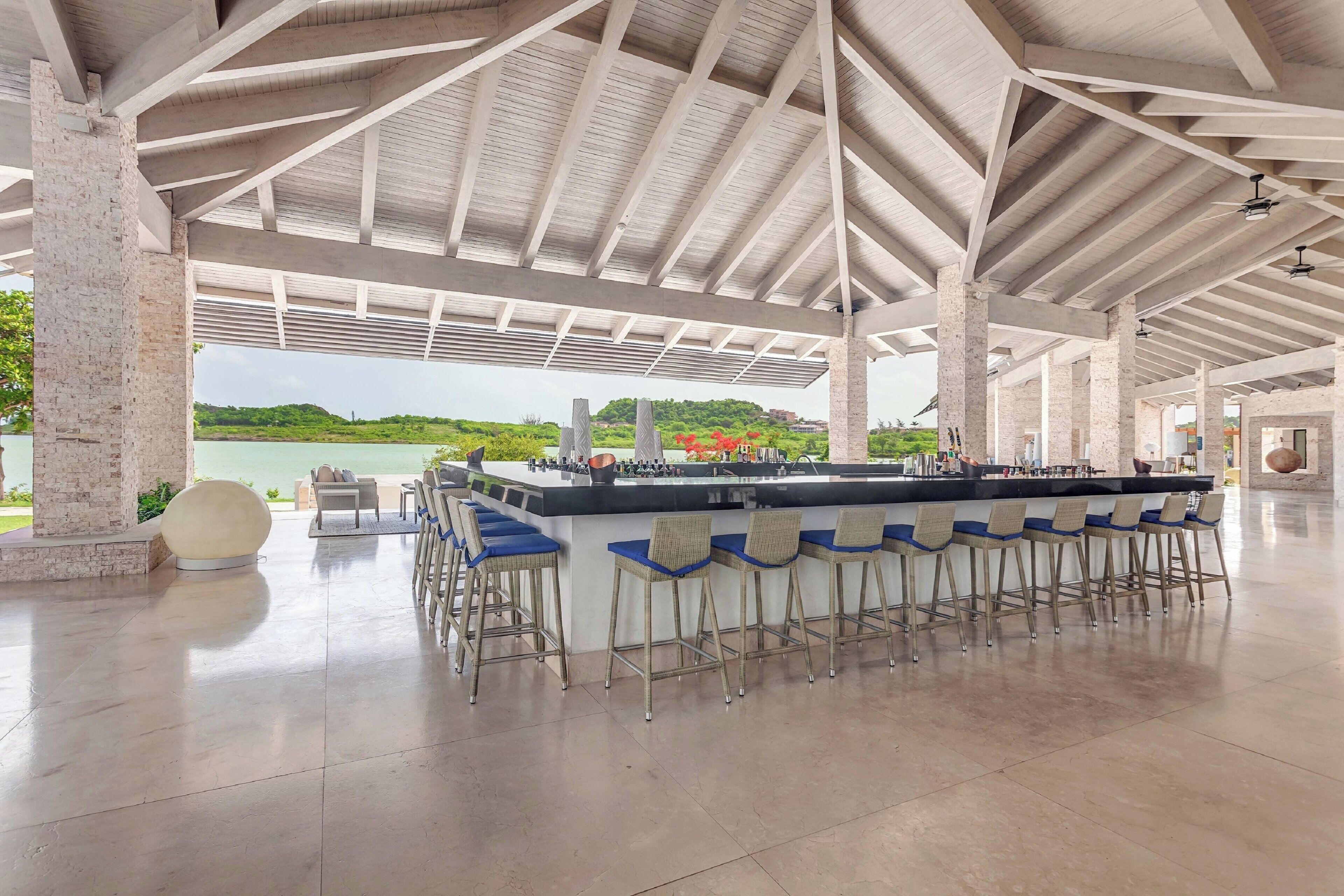 A spacious outdoor bar area with a long counter and multiple bar stools, a large round table, and a view of a natural landscape.