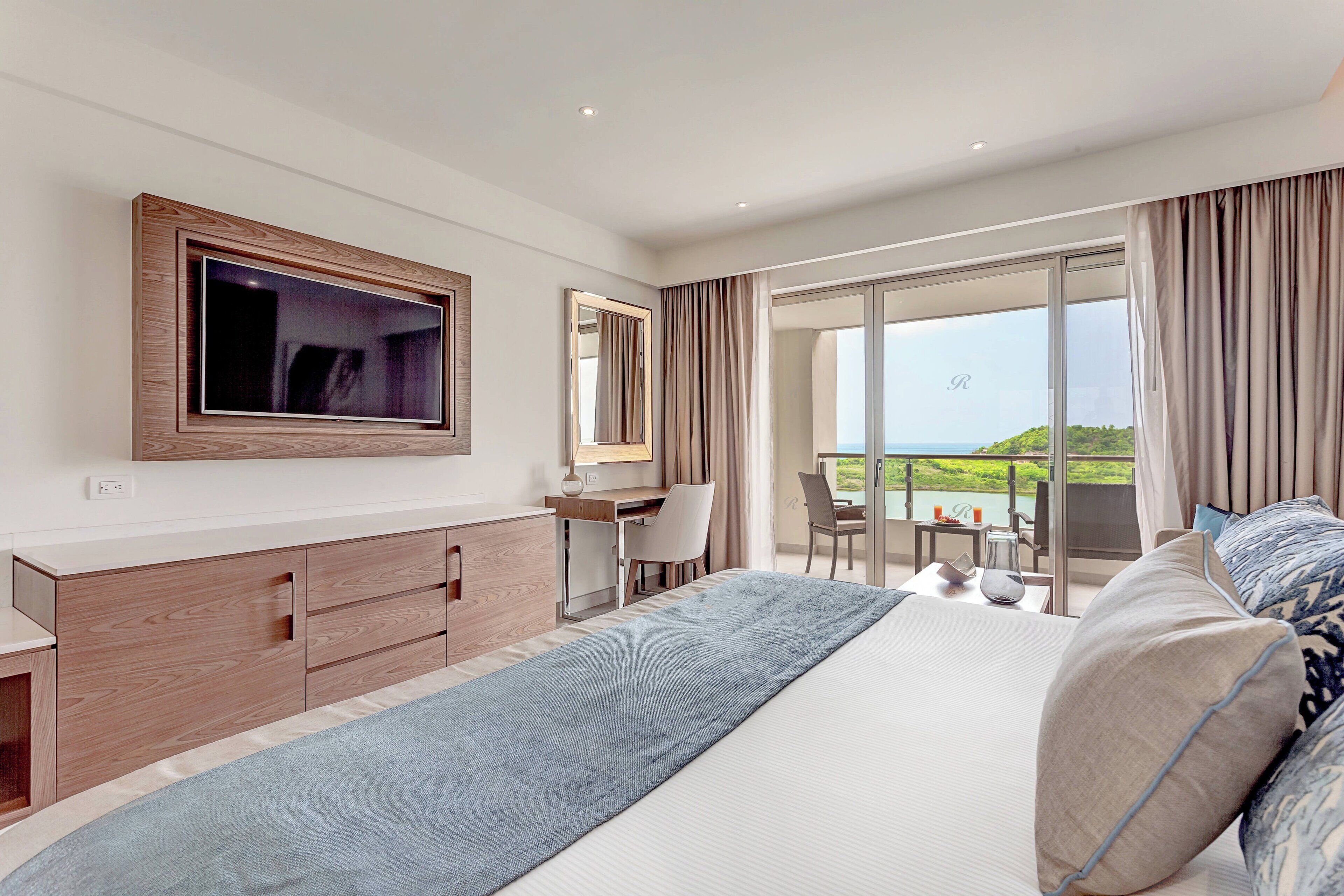 A modern hotel room with a large bed, a desk, a TV, and a balcony with a view.