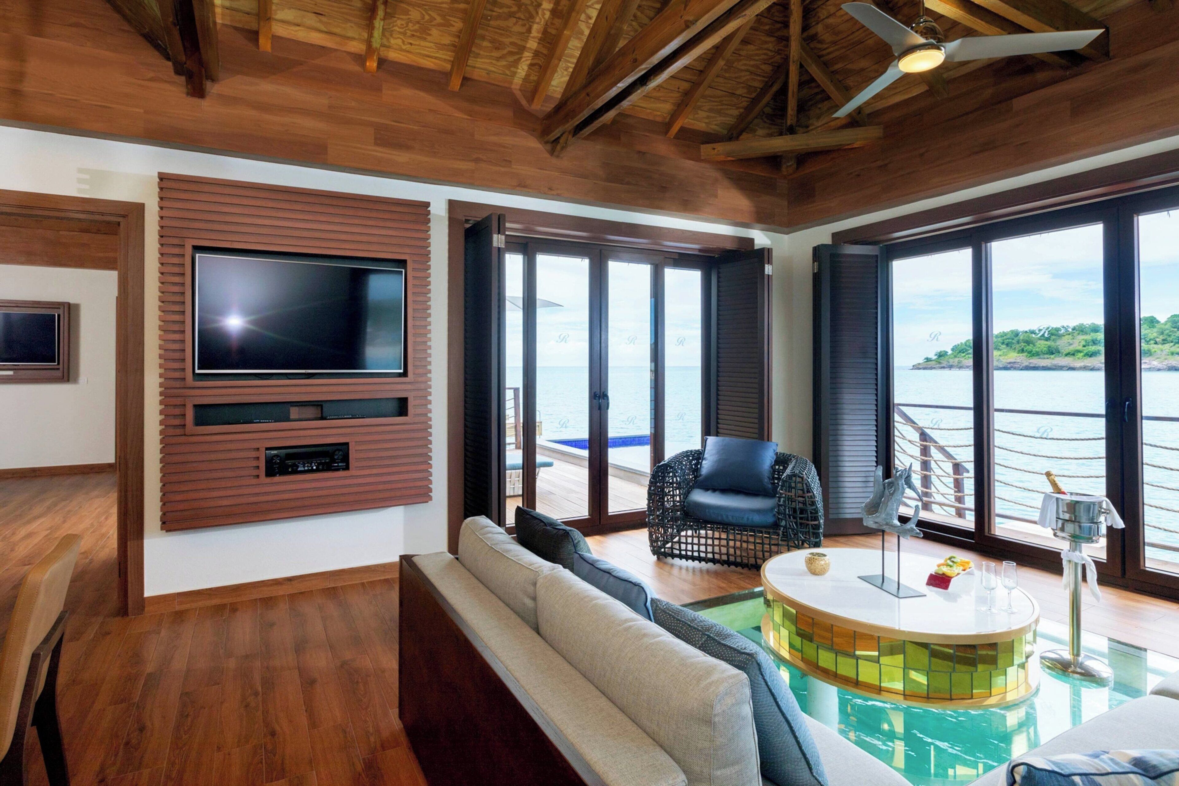 A spacious living area with a large TV mounted on a wooden panel, a comfortable sofa, a black wicker chair, a small pool table, and a view of the ocean through large windows.