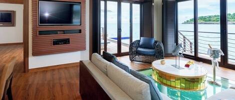 A spacious living area with a large TV mounted on a wooden panel, a comfortable sofa, a black wicker chair, a small pool table, and a view of the ocean through large windows.