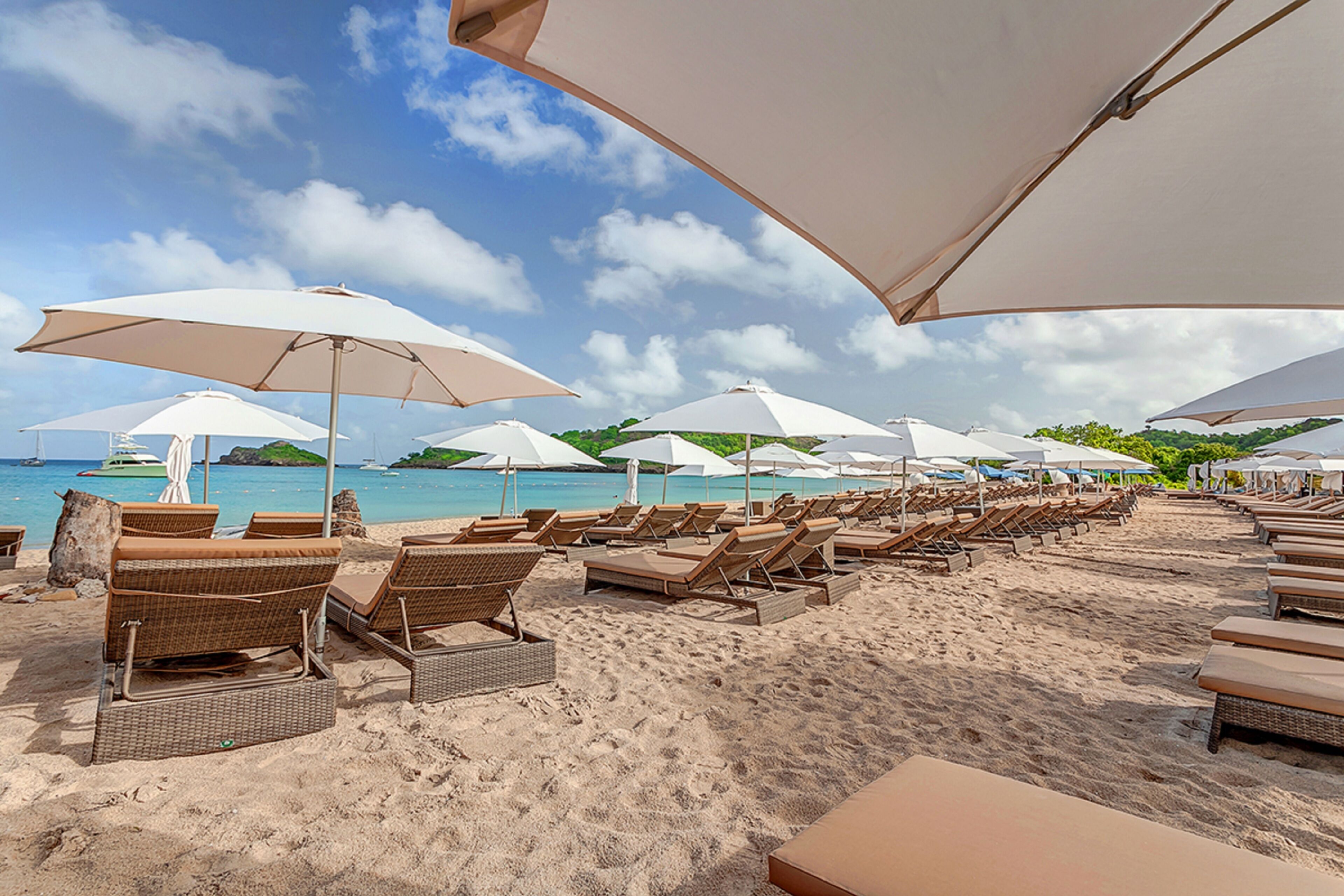 A beach with rows of sun loungers and large umbrellas, a sandy shore, and a view of the sea and distant island.