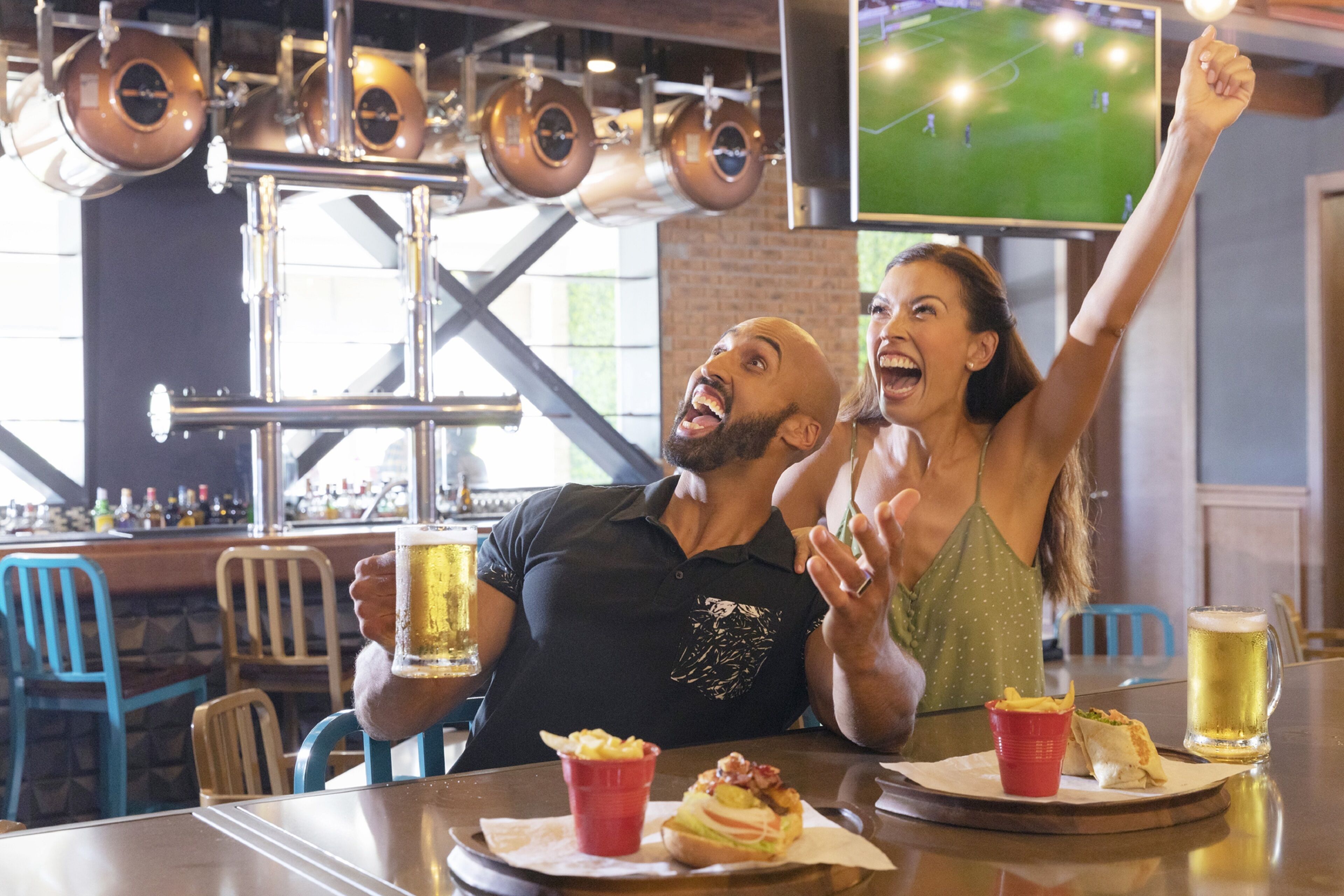 A bar setting with a television displaying a sports game, a man and a woman celebrating, and food and drinks on the table.