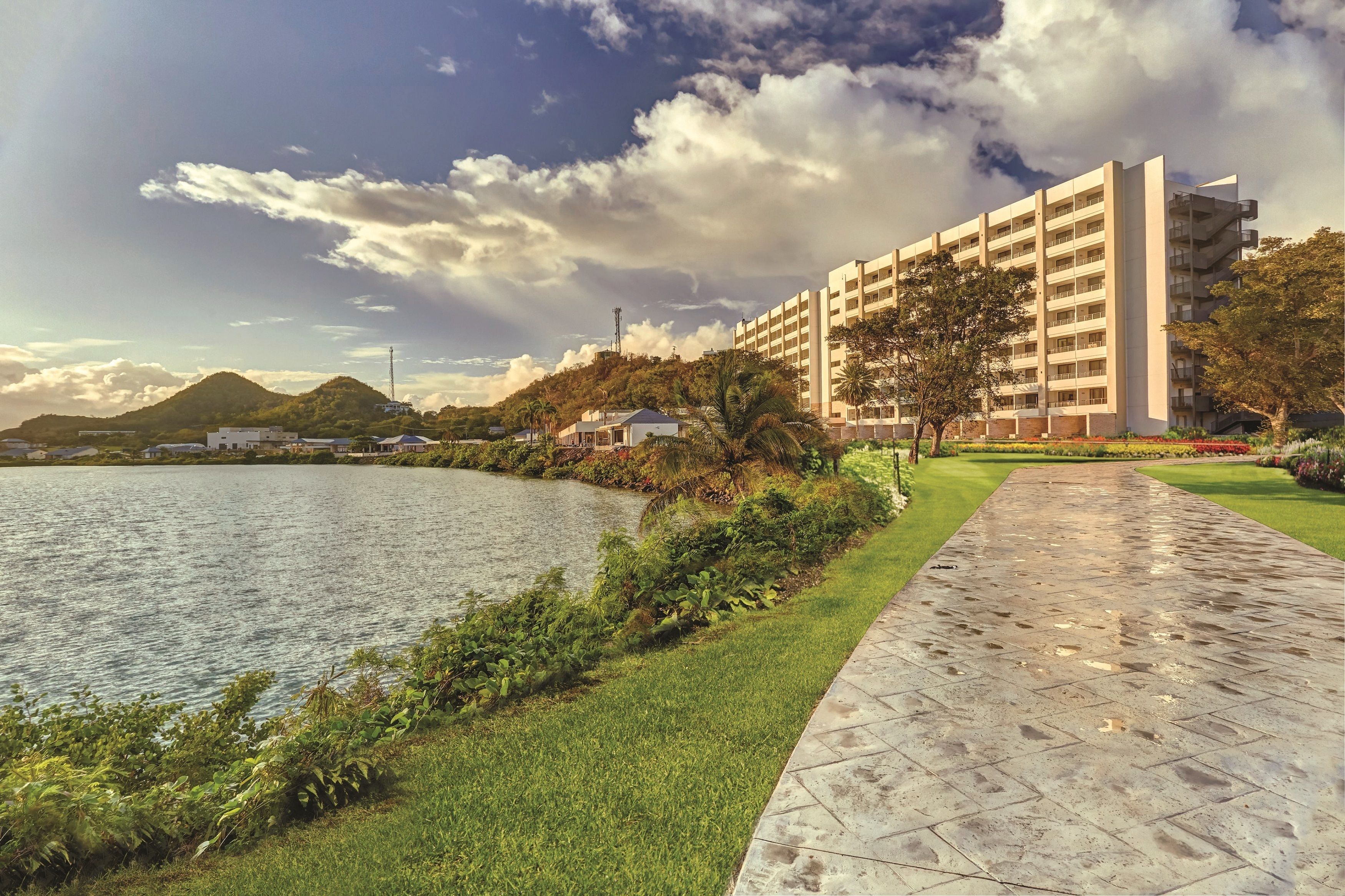 Ein Hotel am Wasserufer mit einem gepflasterten Weg, Grünflächen und einem Berg im Hintergrund.