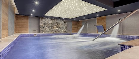 A modern indoor pool with a blue tiled edge, a ceiling with a geometric light fixture, and a wall with a marble-like texture.
