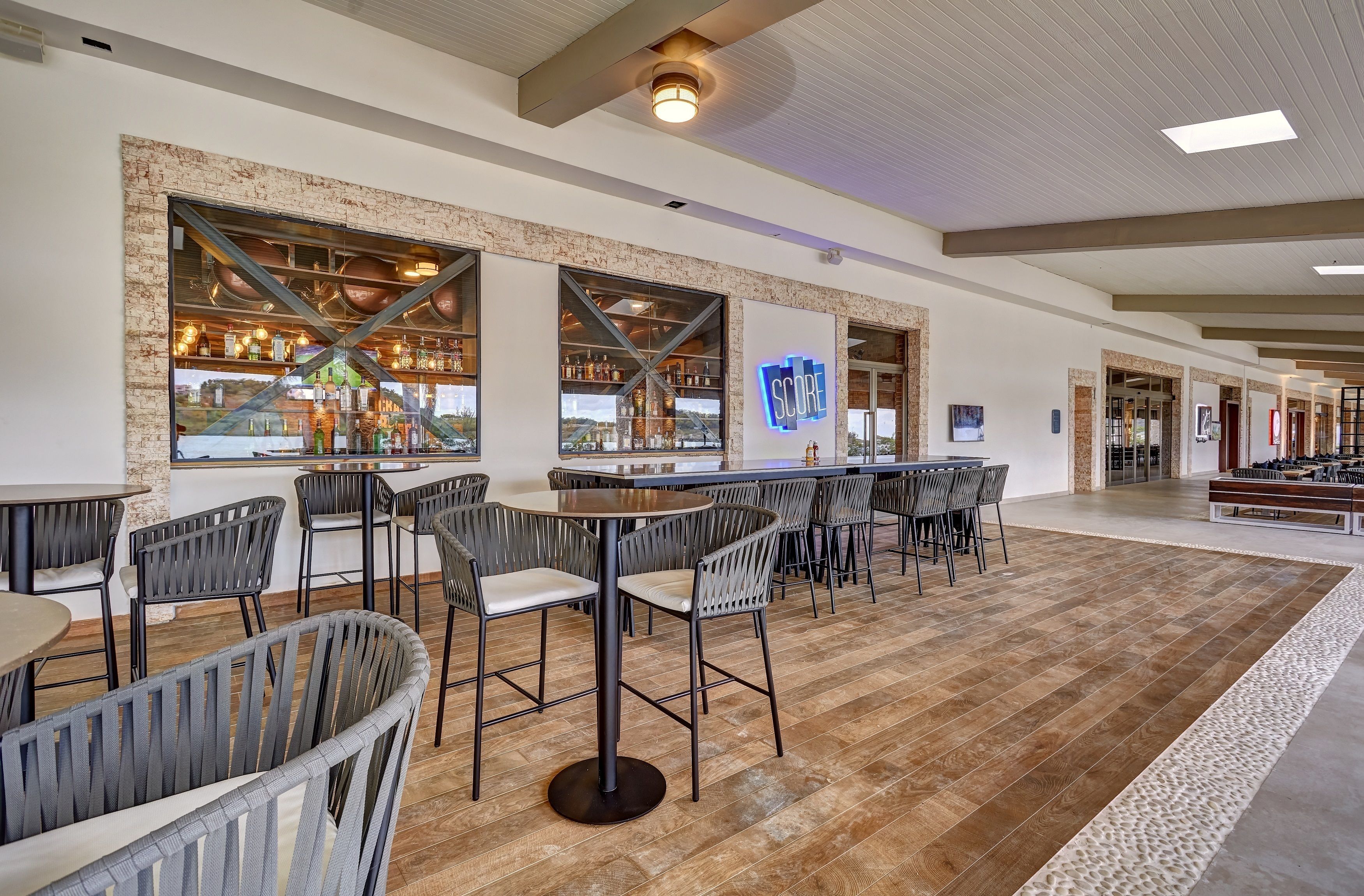 A modern bar area with exposed brick walls, metal beams, and a large illuminated logo. There are multiple seating areas with round tables and chairs, and a wooden floor.